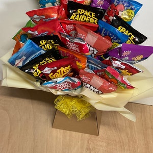 May include: A bouquet of various snack chip bags, including brands like Space Raiders, Frazzles, and Fish 'n' Chips. The bags are arranged in a decorative display, with a yellow bow and a brown box at the base. The chips are in colorful packaging.