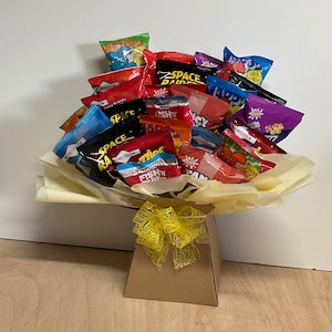 May include: A gift bouquet of various snack-sized chip bags, arranged in a paper-lined box and tied with a yellow ribbon. The chip bags are in a variety of colors, including red, blue, and green, with various brand names visible.
