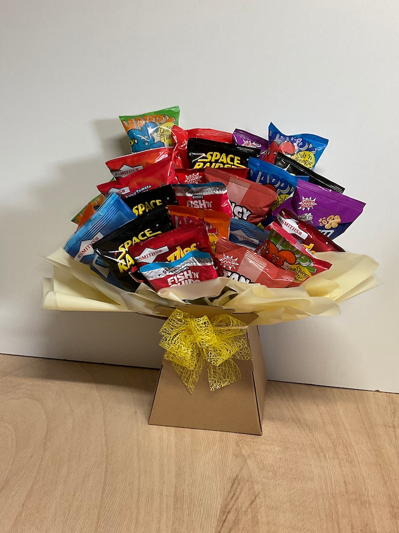 May include: A gift bouquet of various snack-sized chip bags, arranged in a paper-lined box and tied with a yellow ribbon. The chip bags are in a variety of colors, including red, blue, and green, with various brand names visible.