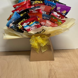 May include: A gift basket filled with various snack-sized chip bags, arranged to resemble a bouquet. The bags feature colorful packaging with text like "Space Raiders" and "Fish 'n' Chips". The basket is brown with a yellow bow.