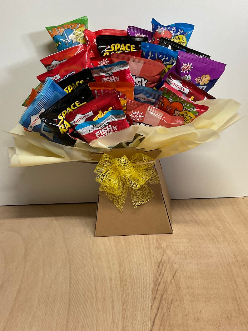May include: A gift basket filled with various snack-sized chip bags, arranged to resemble a bouquet. The bags feature colorful packaging with text like "Space Raiders" and "Fish 'n' Chips". The basket is brown with a yellow bow.