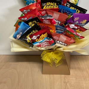 May include: A snack bouquet featuring a variety of colorful chip bags, including "Space Raiders" and "Fish 'n' Chips." The arrangement is presented in a brown box with a yellow ribbon, ideal for gifting. The chips are arranged on a bed of yellow tissue paper.