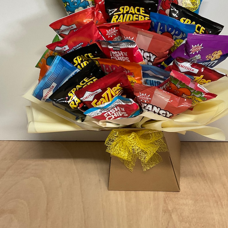 May include: A snack bouquet featuring a variety of colorful chip bags, including "Space Raiders" and "Fish 'n' Chips." The arrangement is presented in a brown box with a yellow ribbon, ideal for gifting. The chips are arranged on a bed of yellow tissue paper.