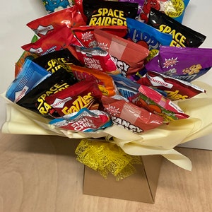 May include: A bouquet of various snack chip bags in a decorative arrangement. The colorful bags include brands like Space Raiders, Frazzles, and Smiths. The arrangement is presented in a yellow tissue paper and a brown box, tied with a yellow ribbon.