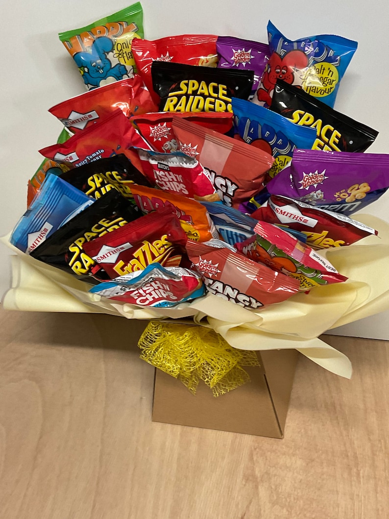 May include: A bouquet of various snack chip bags in a decorative arrangement. The colorful bags include brands like Space Raiders, Frazzles, and Smiths. The arrangement is presented in a yellow tissue paper and a brown box, tied with a yellow ribbon.
