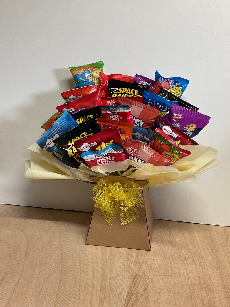 May include: A gift bouquet of various snack chip bags, arranged in a paper-lined box and tied with a yellow ribbon. The chip bags are colorful, with text and graphics, and include brands like Space Raiders and Smiths.