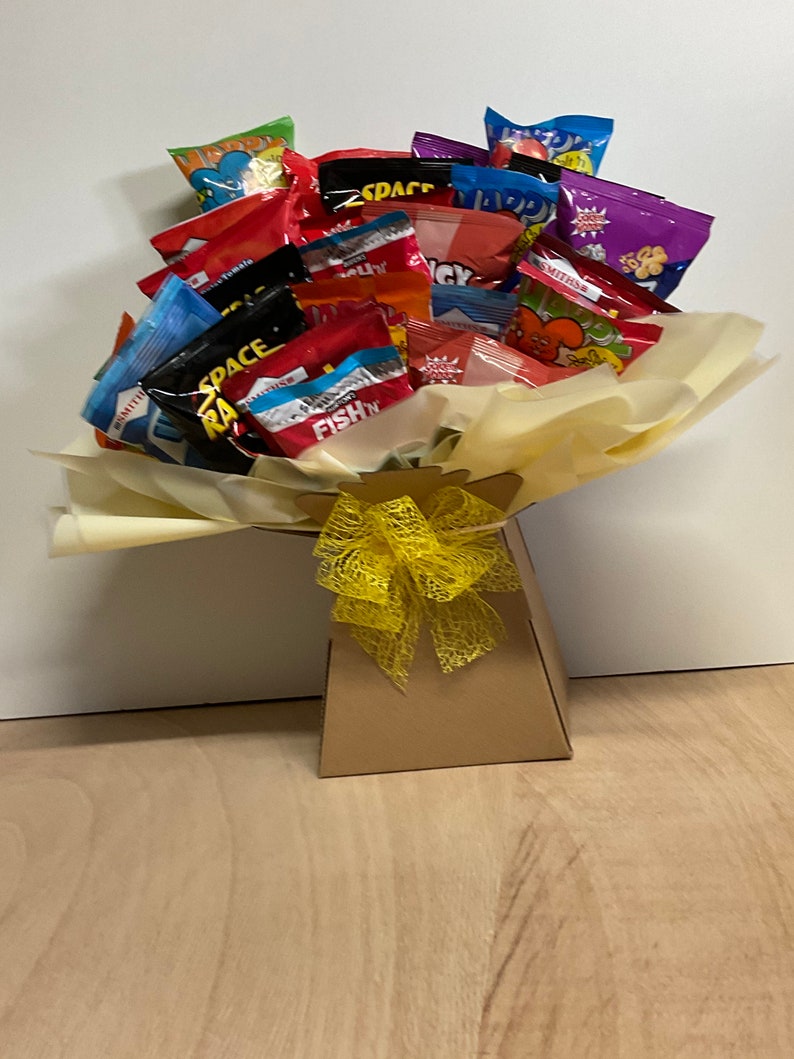 May include: A bouquet of assorted snack bags, including brands like Space Raiders and Smiths, arranged in a brown paper gift bag. The bouquet is wrapped in yellow tissue paper and tied with a yellow ribbon.