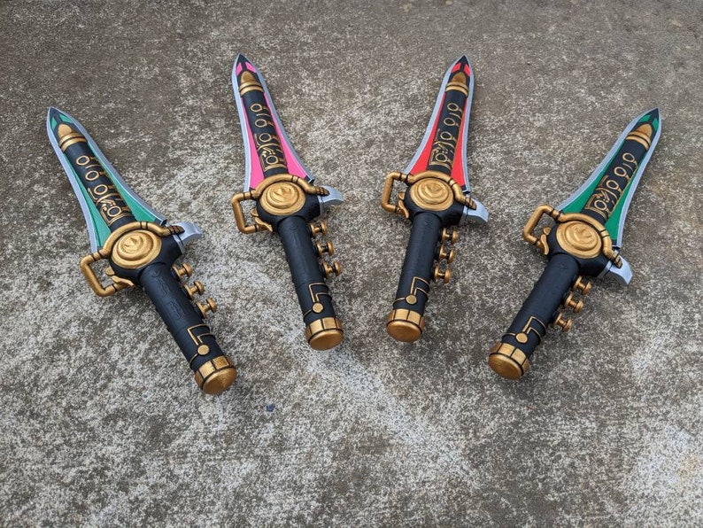 Puede incluir: Cuatro espadas de juguete de pl&aacute;stico negro con detalles dorados y hojas de diferentes colores. Las hojas son verdes, rosas, rojas y verdes. Las espadas tienen la palabra "Power Rangers" escrita en ellas.