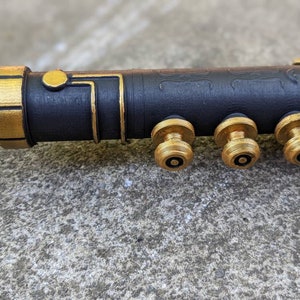 Puede incluir: Un accesorio de metal negro y dorado con tres pomos dorados. El accesorio tiene una superficie texturizada y una banda dorada alrededor del extremo.