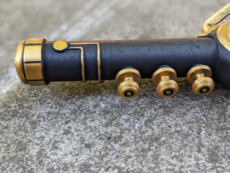 Puede incluir: Un accesorio de metal negro y dorado con tres pomos dorados. El accesorio tiene una superficie texturizada y una banda dorada alrededor del extremo.