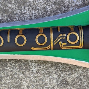Puede incluir: Primer plano de una espada negra y verde con detalles dorados. La espada tiene un dise&ntilde;o decorativo con c&iacute;rculos y l&iacute;neas.