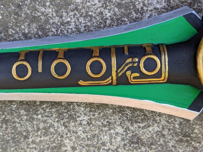 Puede incluir: Primer plano de una espada negra y verde con detalles dorados. La espada tiene un dise&ntilde;o decorativo con c&iacute;rculos y l&iacute;neas.