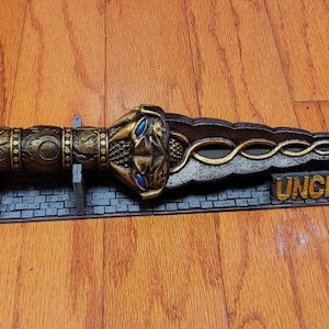 May include: A gold and silver dagger with a detailed head and a snake design on the blade. The dagger is on a display stand with the word "Uncharted" printed on it.