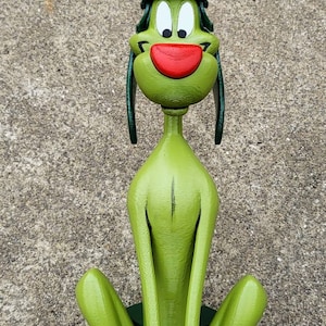 Puede incluir: Una figurita de perro de dibujos animados verde y blanca que lleva un casco verde con la parte superior dorada. El perro está sentado con las piernas cruzadas y tiene la nariz roja y los ojos grandes.