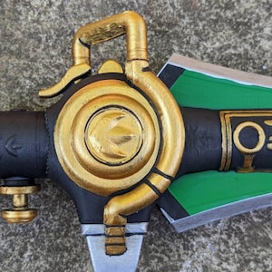 Puede incluir: Primer plano de una empu&ntilde;adura de espada pintada de oro con una hoja negra y verde. La empu&ntilde;adura tiene un dise&ntilde;o circular con un anillo elevado y una peque&ntilde;a palanca. La hoja es parcialmente visible y tiene un esquema de pintura verde y negro.
