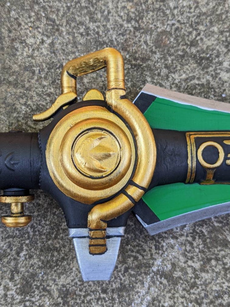 Puede incluir: Primer plano de una empu&ntilde;adura de espada pintada de oro con una hoja negra y verde. La empu&ntilde;adura tiene un dise&ntilde;o circular con un anillo elevado y una peque&ntilde;a palanca. La hoja es parcialmente visible y tiene un esquema de pintura verde y negro.