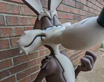 May include: A brown and white cartoon coyote statue with a mischievous expression. The coyote is standing with its right arm raised and its left arm bent at the elbow. The statue is made of a textured material and has a realistic appearance.