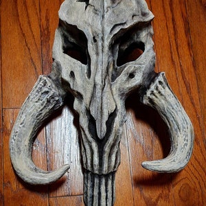May include: A detailed, gray and white, sculpted skull mask with two large horns. The mask has a textured surface and appears to be made of a hard material.
