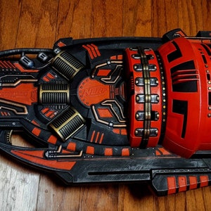 May include: A black and red Nerf blaster with a large, cylindrical magazine. The blaster has a futuristic design with intricate details and the words "The Judge" printed on the side.