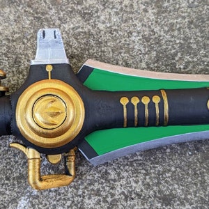 Puede incluir: Una espada negra y verde con detalles dorados. La espada tiene un pomo redondo de color dorado en el mango y una hoja verde con un borde plateado.