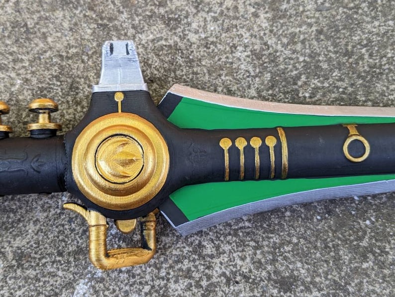 Puede incluir: Una espada negra y verde con detalles dorados. La espada tiene un pomo redondo de color dorado en el mango y una hoja verde con un borde plateado.