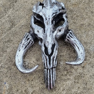 May include: A silver-colored Mandalorian helmet with a skull-like design. The helmet has two horns on the sides and a long, pointed snout.
