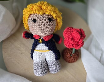 May include: A crocheted plush toy of The Little Prince, with yellow hair, a blue jacket, white pants, and gray boots. The toy is holding a red crocheted rose.