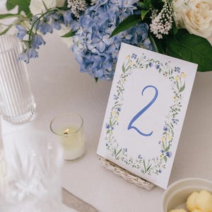 May include: A table setting with a floral centerpiece of white roses, blue hydrangeas, and greenery. A table number card with the number 2 is displayed. Additional elements include glassware, a candle, and a small bowl of butter.