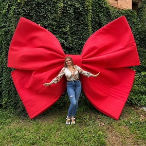 Puede incluir: Un gran lazo decorativo de tela en color rojo brillante, con bucles anchos y estructurados y un nudo central. El lazo está colocado contra una pared cubierta de hiedra verde densa, creando un contraste llamativo entre el rojo y el follaje.