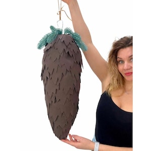 May include: A large, brown paper pinecone decoration with a textured surface. The pinecone is topped with green faux pine needles and suspended by a thin, tan string. A unique, handcrafted holiday or seasonal decoration.