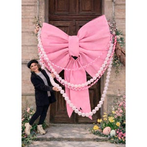 May include: A large, pink bow decoration adorns a wooden door, accented with a pearl garland. The bow is the focal point, with a woman standing nearby. Floral arrangements and Easter eggs add to the festive scene.