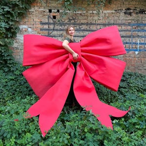 Puede incluir: Un lazo decorativo de color rojo brillante de gran tamaño, fabricado con un material liso y mate. El lazo presenta dos bucles grandes y anchos y dos cintas largas y fluidas que se extienden hacia abajo, creando una pieza visual llamativa.