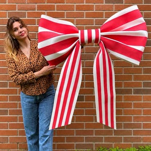 May include: A large, decorative bow with red and white stripes. The bow is held in front of a brick wall and has long, striped streamers. The bow is a festive decoration, possibly for a party or event.