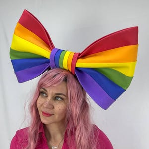 Peut inclure: Un grand serre-tête avec un nœud en forme d'arc-en-ciel. L'arc présente les couleurs de l'arc-en-ciel : rouge, orange, jaune, vert, bleu et violet. L'accessoire est festif.