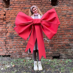 Puede incluir: Un gran lazo rojo brillante con largas cintas. El lazo se sostiene frente a una persona de pie frente a una pared de ladrillos. El lazo es el punto focal de la imagen.