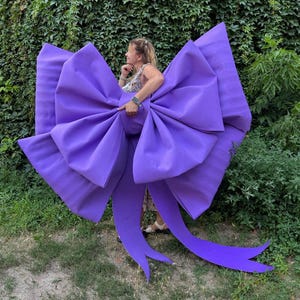 May include: A large, vibrant purple bow with long, flowing tails. The bow is made of a smooth, solid material and is the focal point of the image. The bow is large enough to be worn or used as a decorative element.