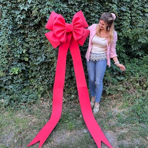 May include: A large, bright red bow with long, flowing ribbons. The bow is made of a sturdy material and is positioned in front of a wall of green foliage. The bow is a festive decoration, suitable for events or celebrations.