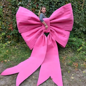 May include: A large, vibrant pink bow, standing upright on the grass. The bow is made of a smooth, possibly fabric material, with long, flowing tails. The bow is large enough for a person to stand behind it. Green foliage forms the background.
