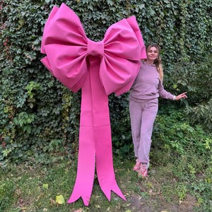May include: A large, vibrant pink bow with long, flowing ribbons. The bow is made of a smooth, possibly fabric material. The bow is standing upright in a grassy area with a green leafy background.