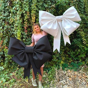 May include: Two large oversized bows, one black and one white, are displayed against a green ivy wall. A person is standing between the bows.