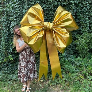 May include: A large, gold bow with long, pointed streamers. The bow is made of a shiny, metallic material and is positioned in front of a wall of green foliage. The bow is a decorative element, possibly for a gift or event.