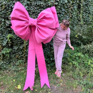 May include: A large, vibrant pink bow with long streamers stands in front of a green leafy background. The bow is made of a smooth material and is a striking decorative element. The bow is large, and the streamers extend downwards.