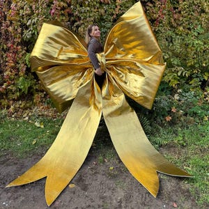 May include: A large, golden bow with long, flowing ribbons. The bow is made of a shiny, metallic material and is positioned outdoors against a backdrop of green and brown foliage. The bow's size suggests it is a decorative element.