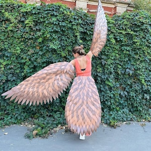 Puede incluir: Una persona con una blusa rosa y un par de alas grandes, marrones y emplumadas. Las alas están hechas de plumas individuales que se superponen y se solapan. La persona está de pie frente a un edificio de ladrillo con hiedra creciendo en las paredes.