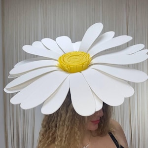 May include: A large, white daisy flower hat with a bright yellow center. The oversized petals are layered, creating a dimensional effect. The hat is a whimsical accessory, perfect for costumes or themed events.
