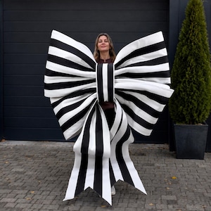 May include: An oversized decorative bow featuring a bold black and white vertical striped pattern. The large bow has two wide, voluminous loops and two long, hanging tails with pointed ends, constructed from a stiff, smooth material.
