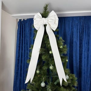 May include: A large white bow sits atop a Christmas tree, with long ribbons cascading down. The tree is decorated with silver and green baubles. Blue curtains form the backdrop.