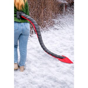 May include: A gray and red devil tail prop, worn around the waist, extends down towards the snow. The tail is gray with red accents and a red tip. The person is wearing a green sweater and blue jeans.