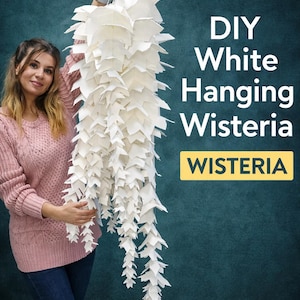 Könnte beinhalten: Eine weiße DIY-Hänge-Wisteria aus Papier, gehalten von einer Person. Die Wisteria besteht aus geschichteten, blattförmigen Papierausschnitten. Der Text auf dem Bild lautet "DIY White Hanging Wisteria" und "WISTERIA".