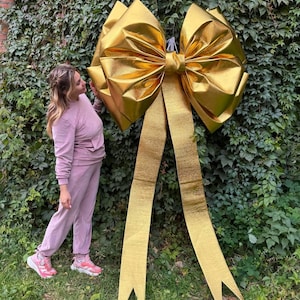 May include: A large, golden bow with long, flowing ribbons. The bow is made of a shiny, metallic material and is displayed against a backdrop of green foliage. The bow is a decorative element, suitable for events or celebrations.